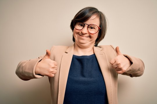 Young down syndrome business woman wearing glasses standing over isolated background success sign doing positive gesture with hand, thumbs up smiling and happy. Cheerful expression and winner gesture.
