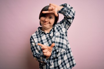 Young down syndrome woman wearing casual shirt over pink background smiling making frame with hands and fingers with happy face. Creativity and photography concept.