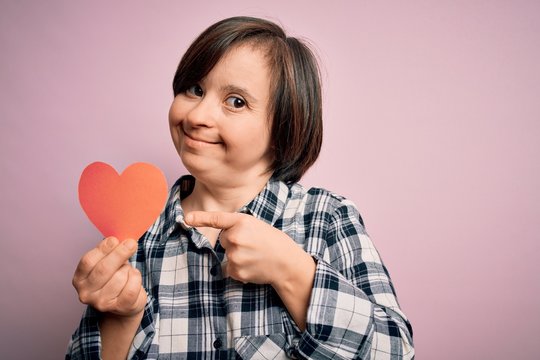 Young down syndrome romantic woman holding red heart paper shape over pink background very happy pointing with hand and finger