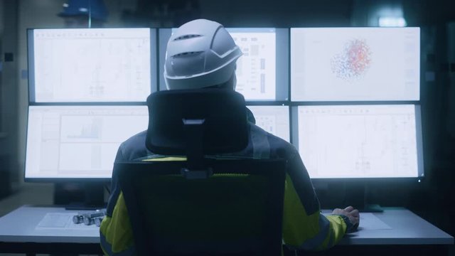 Industry 4.0 Modern Factory: Facility Operator Wearing Hardhat and Safety Vest Controls Workshop Production Line, Uses Computer with Screens Showing Machinery Control Operation UI. Back View Zoom In