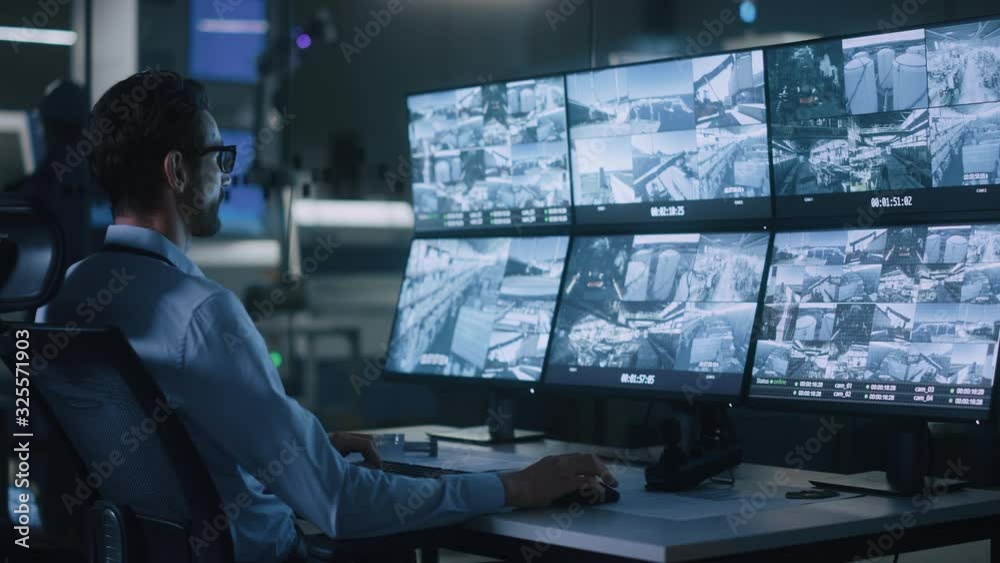 Poster Industry 4.0 Modern Factory: Security Operator Controls Proper Functioning of Workshop Production Line, Uses Computer with Screens Showing Surveillance Camera Footage Feed. High-Tech Security