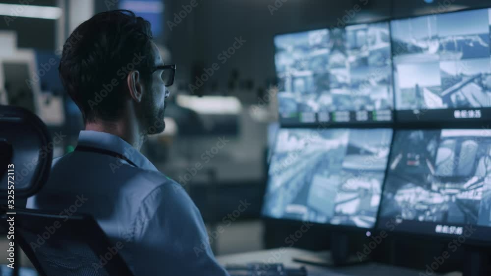 Poster Industry 4.0 Modern Factory: Security Operator Controls Proper Functioning of Workshop Production Line, Uses Computer with Screens Showing Surveillance Camera Footage Feed. High-Tech Security