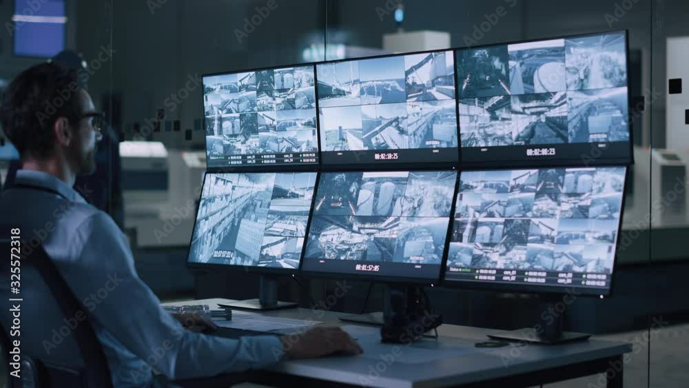 Poster Industry 4.0 Modern Factory: Security Operator Controls Proper Functioning of Workshop Production Line, Uses Computer with Screens Showing Surveillance Camera Footage Feed. High-Tech Security