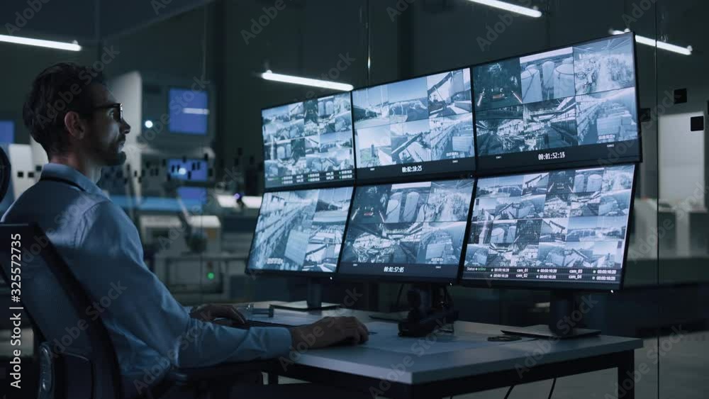Poster Industry 4.0 Modern Factory: Security Operator Controls Proper Functioning of Workshop Production Line, Uses Computer with Screens Showing Surveillance Camera Footage Feed. High-Tech Security