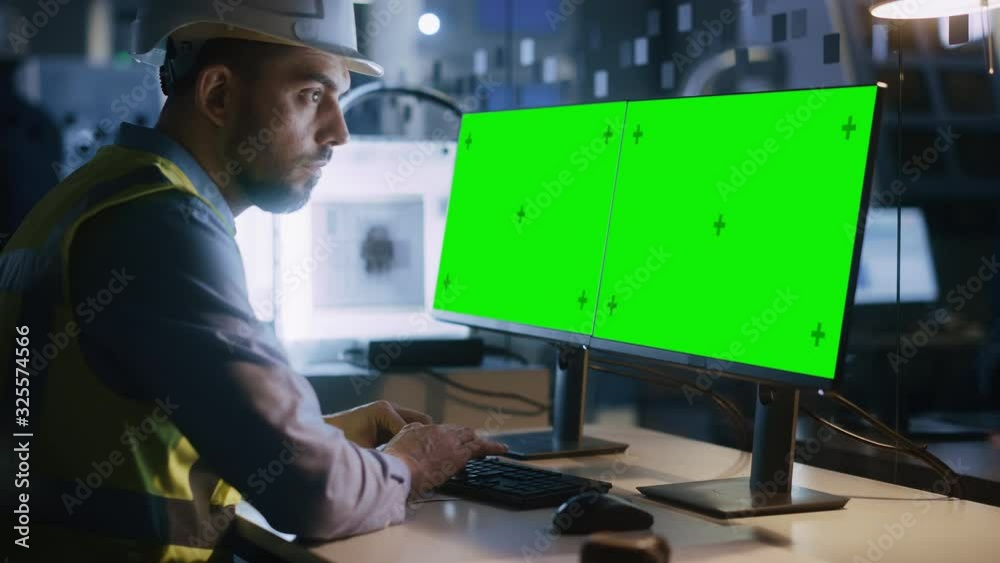 Poster Engineer Wearing Safety Vest and Hardhat Works on a Computer, Two Monitor Screens Show Chroma Key / Green Screen Displays. Modern High-Tech Industry 4.0 Electronics Production Factory