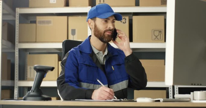 Caucasian man talking on mobile phone while working at post office store with parcels and on computer. Postman registering box and filling in invoice while speaking on cellphone in mail storage.