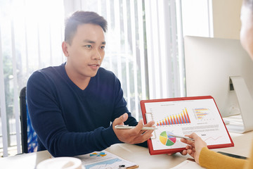 Handsome young Vietnamese businessman showing document with chart and circle diagram to colleague at meeting