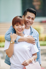 Happy young Asian man hugging his pretty girlfriend with many shopping bags from behind when that are standing outdoors