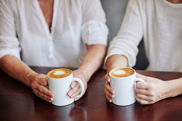 Hands of senior woman and her adult daughter drinking big mugs of cappuccino and talking in cafe