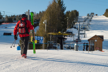 Ski Patrol on Ski Hill walking towards ski chairlift carrying green skis on blue sky day wearing ski patrol uniform with white cross on back horizontal format room for type