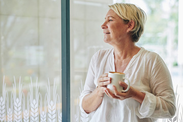 Positive mature woman in white cotton blouse drinking mug of coffee and looking through window