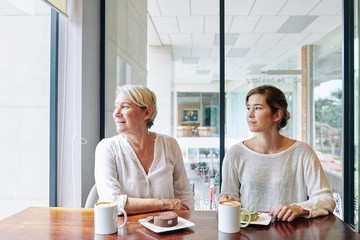 Beautiful mature woman and her adult daughter drinking coffee and eating tasty swiss rolls in local cafe