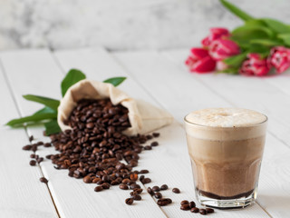 Cappuccino or latte with milk froth in a glass. Сoffee beans in a bag of coffee and coffee leaves on a white wooden background.Spring flowers, tulips in the background. Soft focus, close up.
