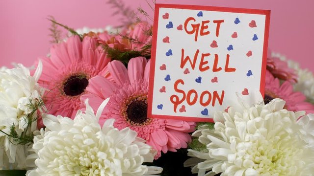 A get well soon message card with a beautiful bouquet of pink and white flowers. Woman hands placing a get well soon wishing card with gerbera daisy flowers and chrysanthemums flowers in the backgr...