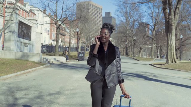 Fashionable Young African American Business Woman Walking Through Park With Luggage While Talking On Phone Traveling On A Sunny Day