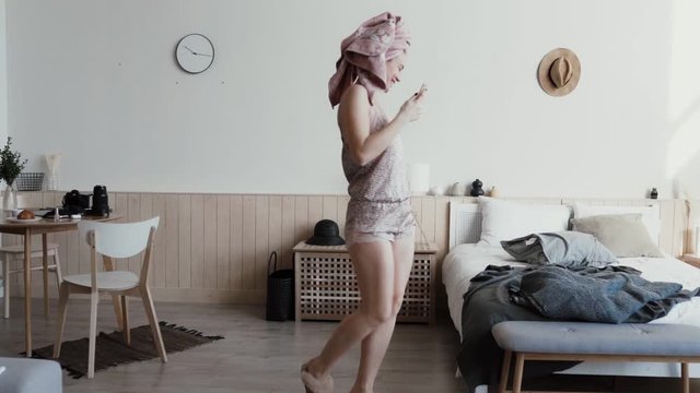 Cheerful woman get out bed throws pillow for joy. Funny girl dressed nightwear and home slippers wearing wireless headphones dancing hair wrapped in bath towel. Morning fun background of accessories
