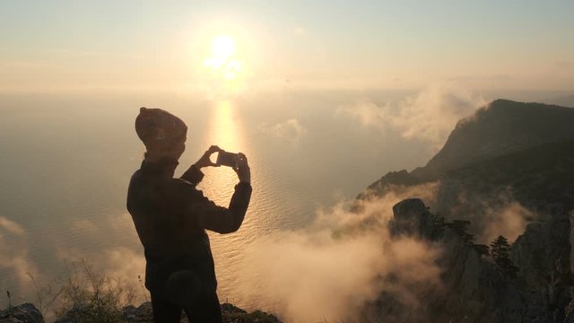 Close-up shooting of a sporty tourist woman wearing a bandana standing on the edge of a high mountain, who photographs a beautiful sunset and cloud smoke and steam over the sea on her smartphone phone