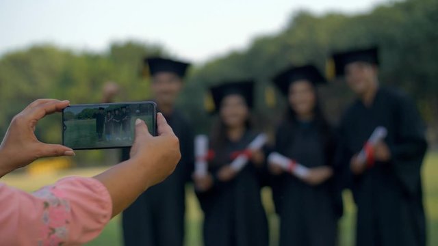 Pan shot - Woman hands clicking pictures of college pupils on graduation day. Happy group of young teens posing for the photos while holding their degrees in graduation gowns  - convocation ceremony