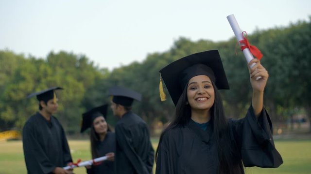 Attractive Indian graduate posing towards the camera with a big smile on her face. Young graduated girl holding her graduation degree in pride while wearing the academic black gown and cap - convoc...