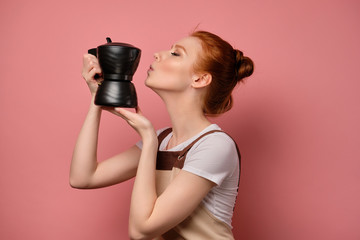 Obraz premium A cute red-haired girl with a high bun in a apron stands in profile on a pink background and reaches for a kiss in a coffee pot