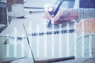Hand taking notes in notepad. Forex chart holograms in front. Concept of research. Double exposure