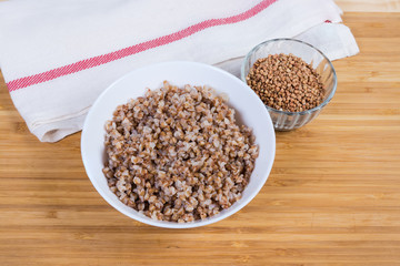 Prepared buckwheat porridge in white bowl and uncooked buckwheat groat