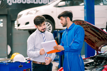 Vehicle maintenance handsome man servicer explain car condition, point at vehicle part to male customer in garage. Automotive mechanic hold automotive checklist document. Car repair service concept.