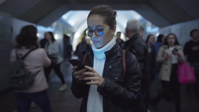 Woman standing in the entrance of a subway station using a smartphone while people walk around her