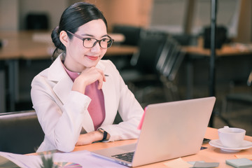 Happy attractive Asian business woman thinking while working on laptop and holding coffee cup with smile in Modern office or living room with copy space