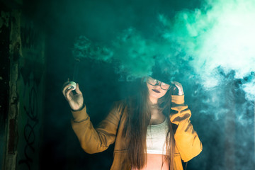 Blonde hair girl holds a smoke bomb that throws green smoke around. Young girl wears an urban style a cap and yellow jacket.