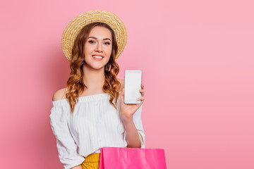 Happy smiling young woman in a straw hat and white dress with shopping bag posing isolated on pink background, copy space. Girl holds smartphone in hands, mockup, empty screen, space for design
