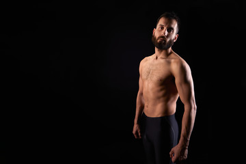 Fototapeta na wymiar Caucasian young man with beard, serious, shirtless, with tights, muscular body, on black background looking straight ahead, horizontal