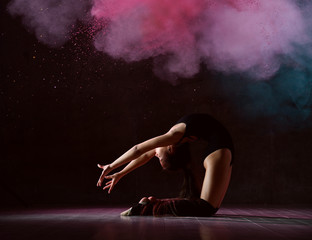 Young girl gymnast in black sport body and special footwear making gymnastic pose over dark background with pink smoke