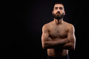 Caucasian young man with a beard, serious, no shirt, muscular body,on black background looking straight ahead with arms crossed, horizontal