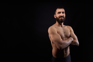 Fototapeta na wymiar Caucasian young man with a beard, smiling, shirtless, muscular body,on black background looking straight ahead with arms crossed, horizontal
