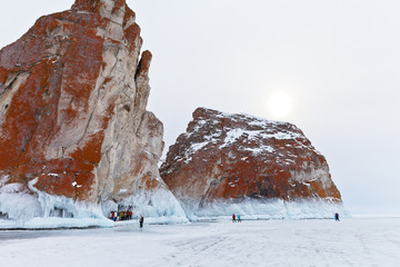 Baikal Lake in the winter. Tourists travel on the ice of frozen lake to the northern edge of Olkhon Island. A group of tourists is photographed against icy rocks of Three Brothers of Cape Sagan-Hushun