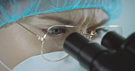 Woman in eyeglasses and hospital cap looking through a microscope