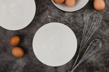 Dark baking background with eggs, whisk, eggshell and white plates. Copy space, top view.