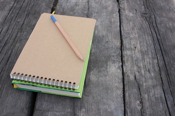 Book, paper notepad on spiral and pencil on rough textured wooden table in park. Top view