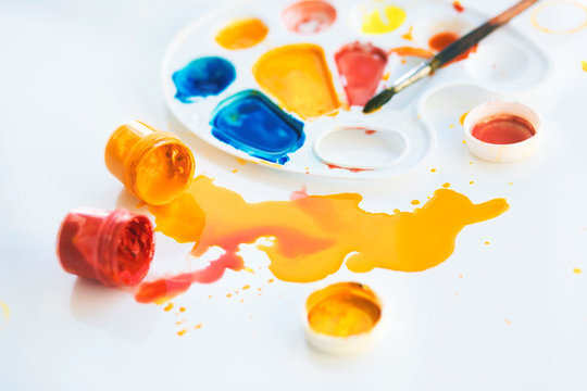 On a white table is a palette on which paints, a brush are mixed, and next to it are overturned jars of gouache, from which the paint flows. - Powered by Adobe