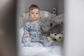 Adorable blond toddler sitting in his crib in the morning and holding his favorite toy.