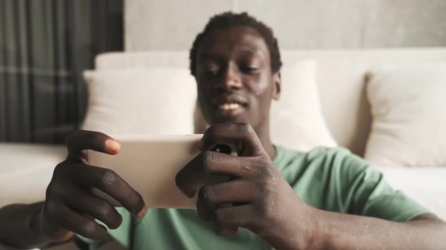 An attractive african american man in casual clothes is using his smartphone while holding it horizontally in the living room