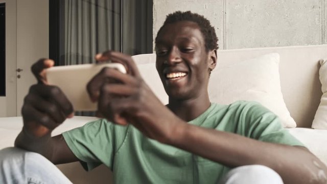 A happy laughed african american man in casual clothes is watching something on his smartphone while holding it horizontally in the living room