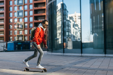 Obraz na płótnie Canvas Stylish hipster in red jacket, sunglasses, sneakers and backpack riding on longboard with one foot placed on board and pushing off with the other. Selective focus. Concept of leisure activity, urban.