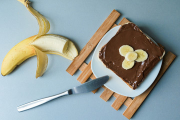 A delicious piece of toast with chocolate spread and banana slices on a modern kitchen countertop with a kitchen knife, plate and vintage wooden surface