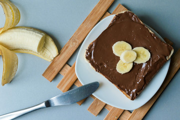 A delicious piece of toast with chocolate spread and banana slices on a modern kitchen countertop with a kitchen knife, plate and vintage wooden surface