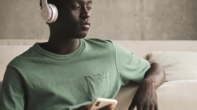 A positive young african american man with wireless headphones is listening to the music while using his smartphone in the living room