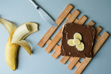 A delicious piece of toast with chocolate spread and banana slices on a modern kitchen countertop with a kitchen knife, plate and vintage wooden surface