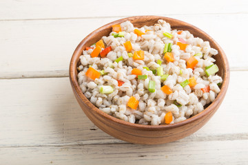 Pearl barley porridge with vegetables in wooden bowl on a white wooden background. Side view, close up.