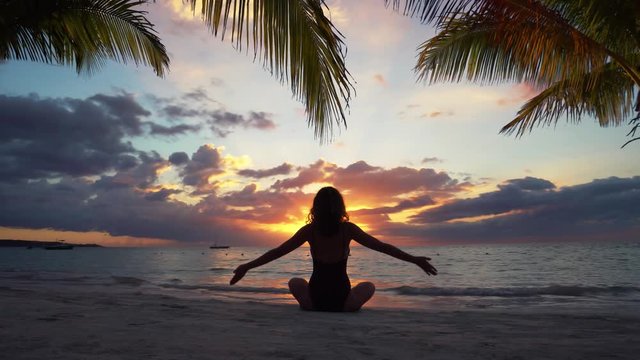 Woman silhouette on sunset making yoga on the beach under tropical palmas 